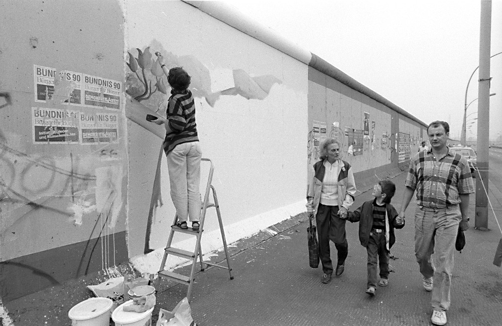 Artist paints artwork at the East Side Gallery, woman, man and child walk by